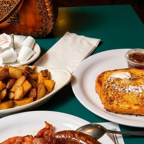 Breakfast spread with French toast, home fries, sausage, bacon, and condiments on a green table.