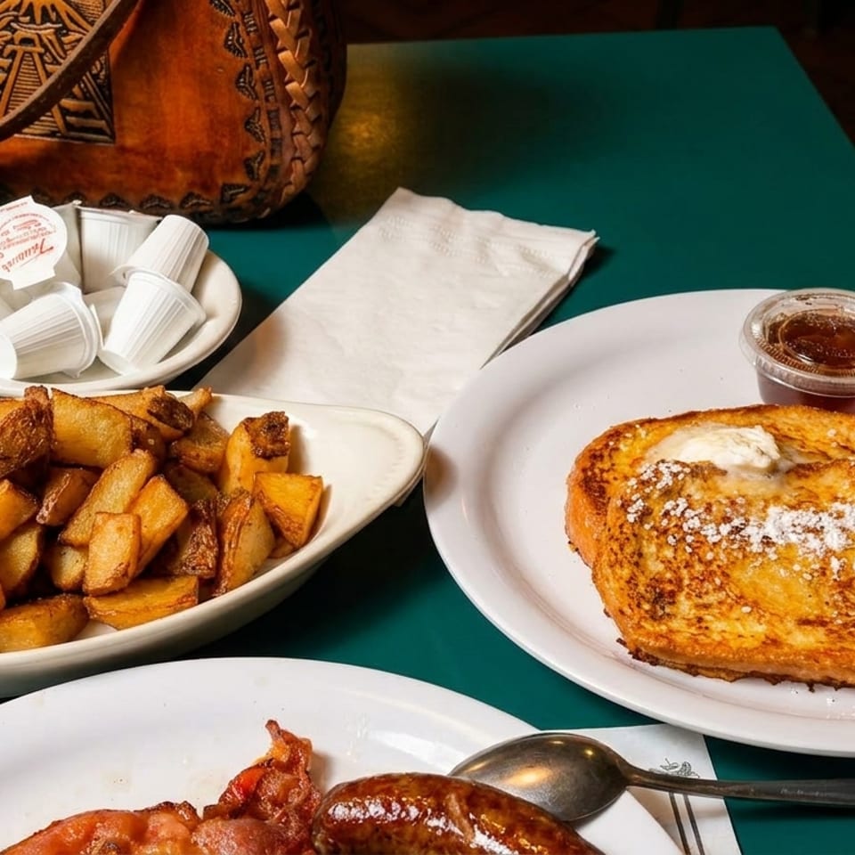 A breakfast spread with French toast, home fries, sausage, bacon, and coffee creamers on a green table.