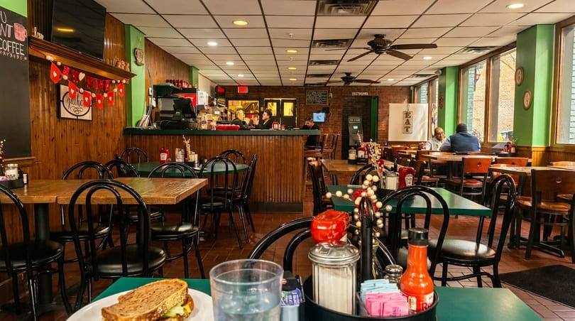 Cozy diner interior with wooden paneling, green walls, patrons dining, and a sandwich on a table.